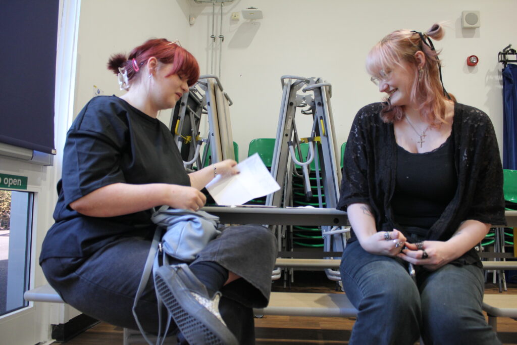 Two female Post-16 students are pictured sat at a table together and reading their A-Level results with smiles on their faces.